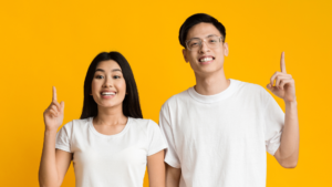 Comment prendre soin de sa vue ? Homme qui porte des lunettes et femme aux cheveux longs, en tshirt blanc, ils lèvent le doigt. Le fond est uni et jaune.