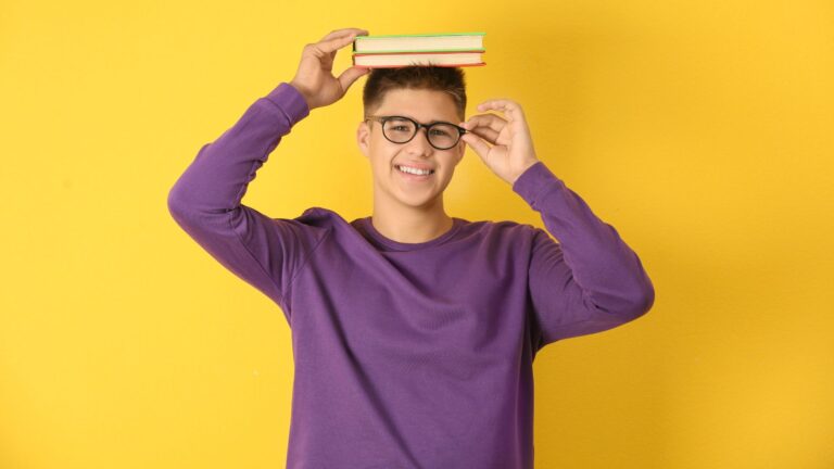 Ados et myopie : fond jaune, jeune homme avec pull violet, des lunettes, des livres tenus en équilibre sur sa tête.